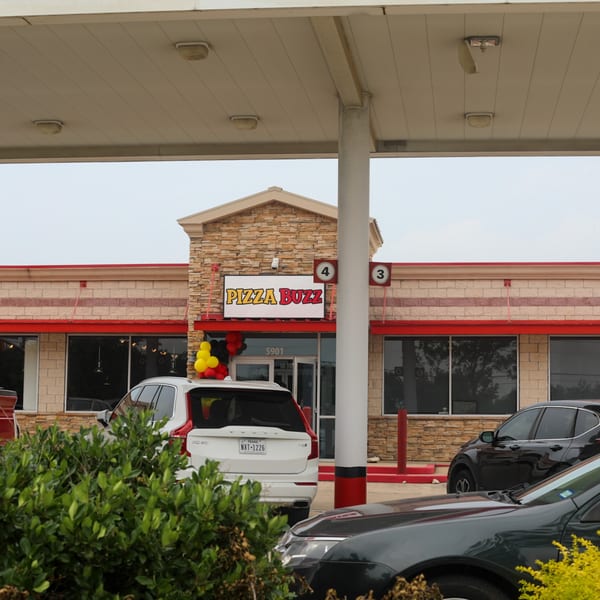 The exterior of Pizza Buzz restaurant with a stone facade, red awnings, and cars parked outside.