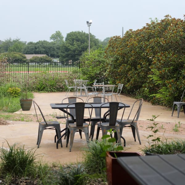 Outdoor patio with several metal tables and chairs, surrounded by lush green foliage.