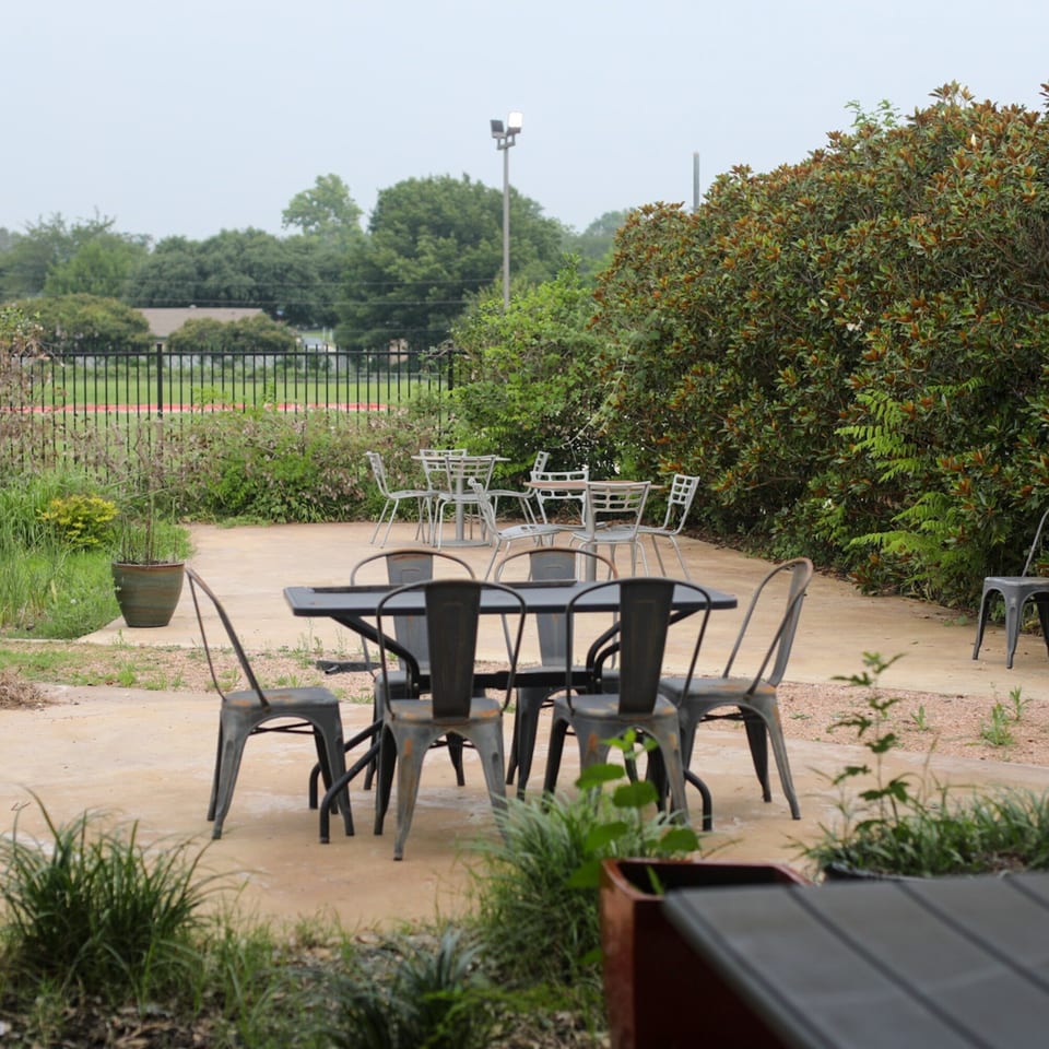 Outdoor patio with several metal tables and chairs, surrounded by lush green foliage.