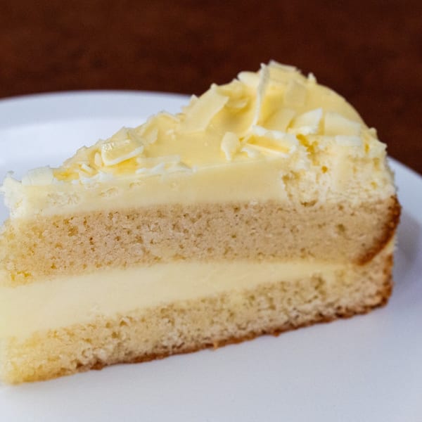 A slice of white cake with creamy white frosting and white chocolate shavings on a white plate.