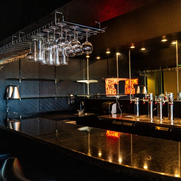 A dark, empty bar at Pizza Buzz with hanging glasses, beer taps, and a neon "Welcome Pizza Lovers" sign.