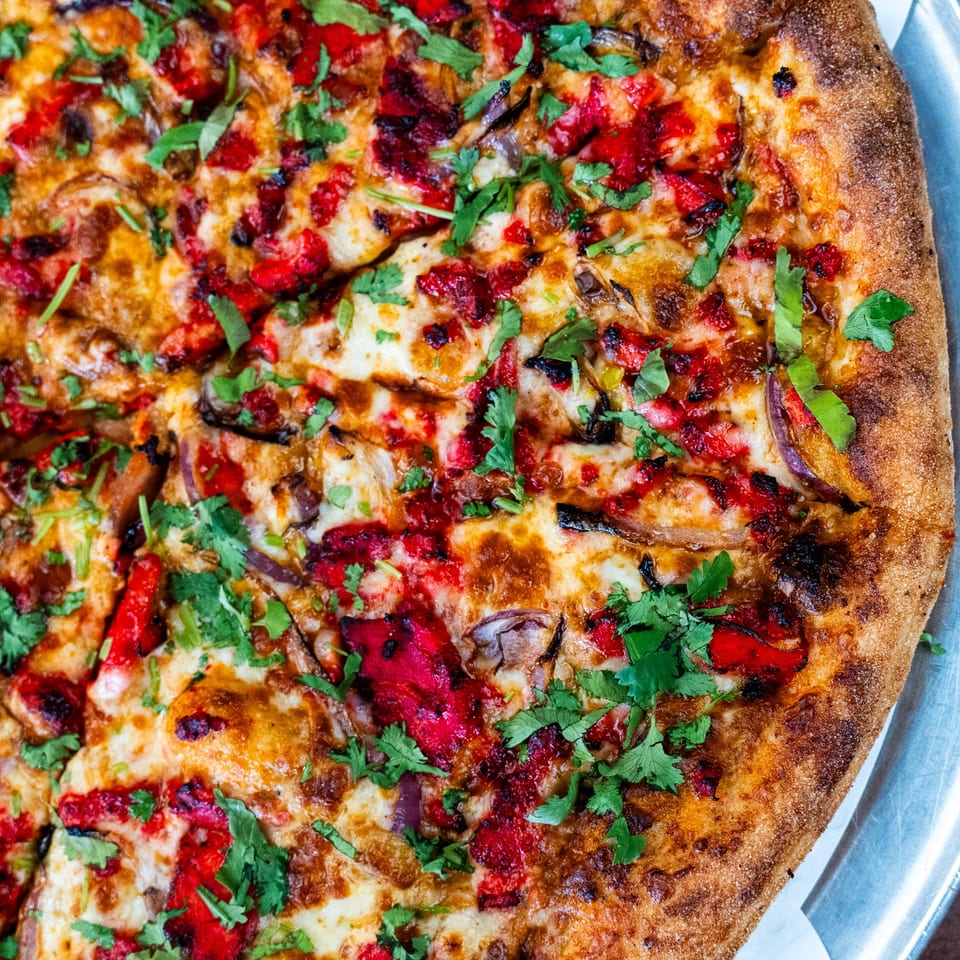 Close-up of a Pizza Buzz pizza with red peppers, onions, and fresh cilantro on a metal tray.