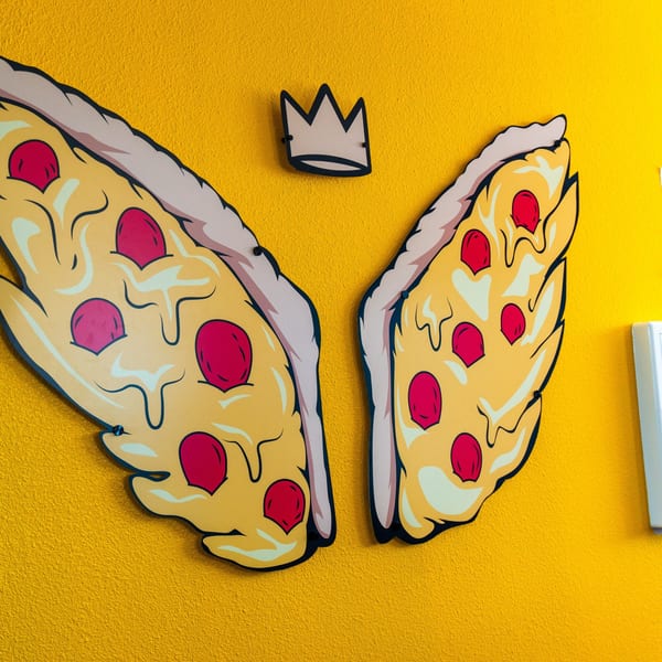 Pizza Buzz wall art features two pizza slices as wings with a crown above on a yellow wall.