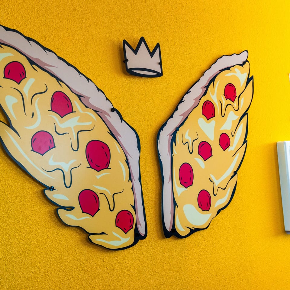 Pizza Buzz wall art features two pizza slices as wings with a crown above on a yellow wall.
