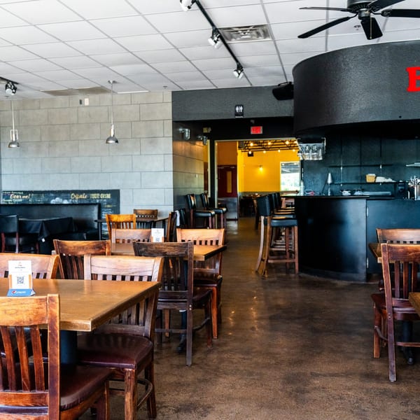 Interior of Pizza Buzz restaurant with wooden tables and chairs, a bar area, and a chalkboard menu.