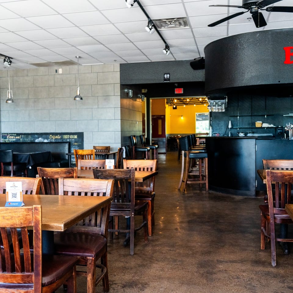 Interior of Pizza Buzz restaurant with wooden tables and chairs, a bar area, and a chalkboard menu.