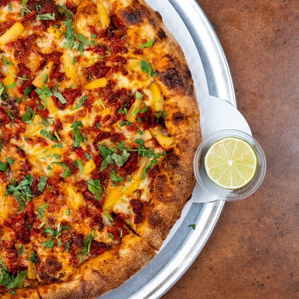 Pizza Buzz pizza with pineapple, red sauce, cheese, and cilantro, with a lime wedge.