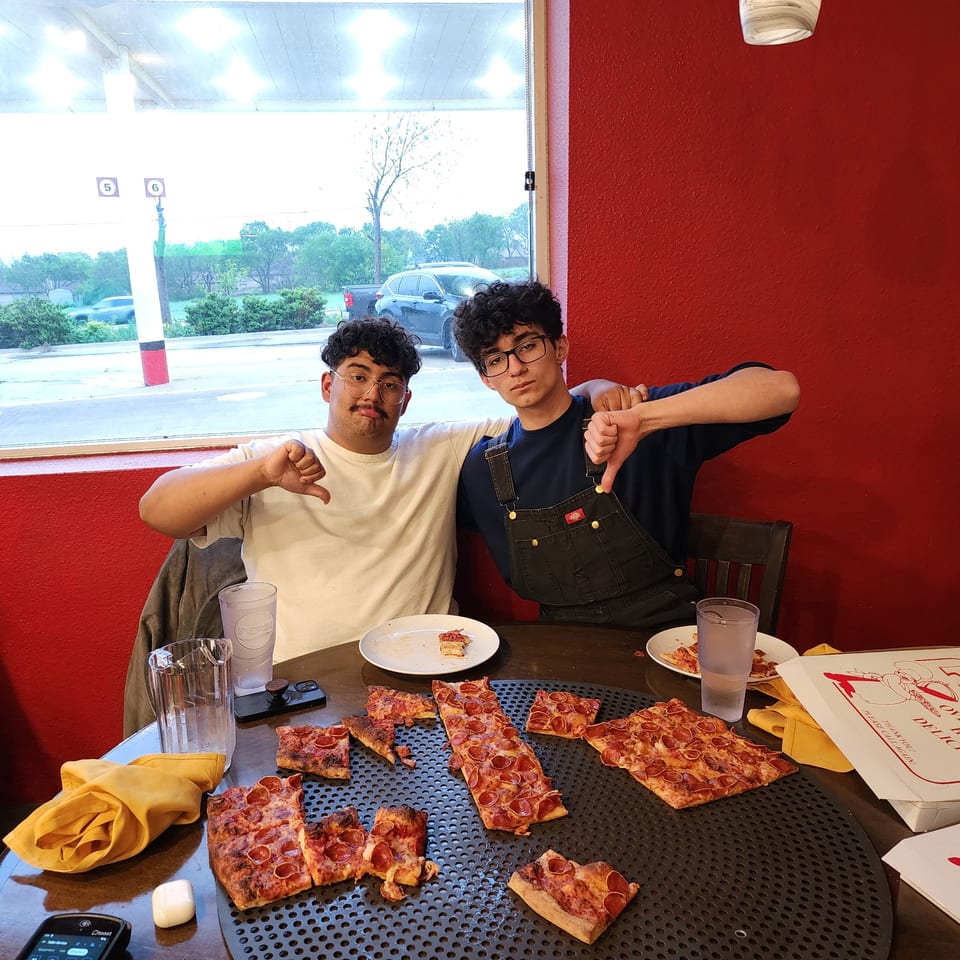Two young men giving thumbs down at Pizza Buzz, surrounded by pepperoni pizza slices.