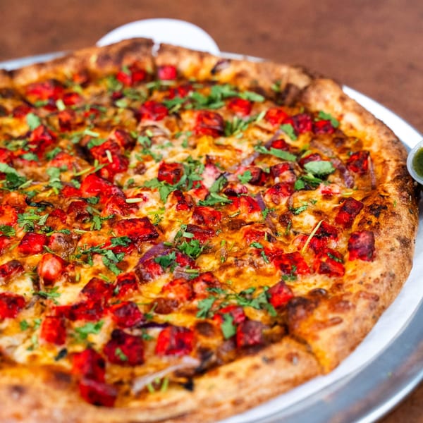 A close-up of a Pizza Buzz pizza, topped with vibrant red chicken tikka, red onions, and fresh cilantro.
