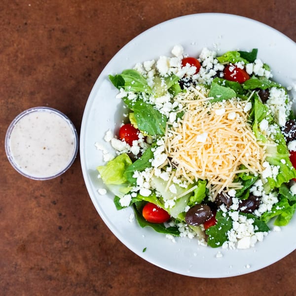 A fresh salad with lettuce, tomatoes, olives, feta, and shredded cheese, with a side of dressing.