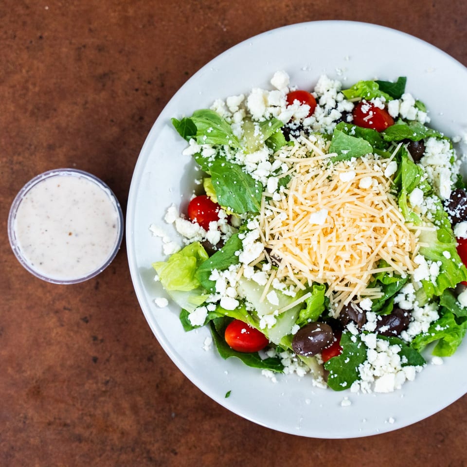 A fresh salad with lettuce, tomatoes, olives, feta, and shredded cheese, with a side of dressing.