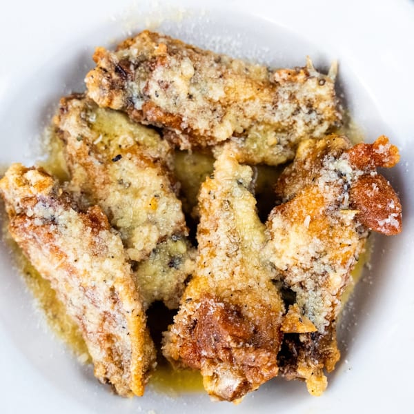 Close-up of crispy, golden-brown chicken wings coated in Parmesan cheese and herbs in a white bowl.