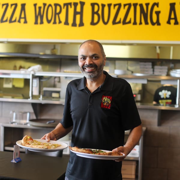 A smiling Pizza Buzz employee holds two plates of pizza slices in a restaurant kitchen.