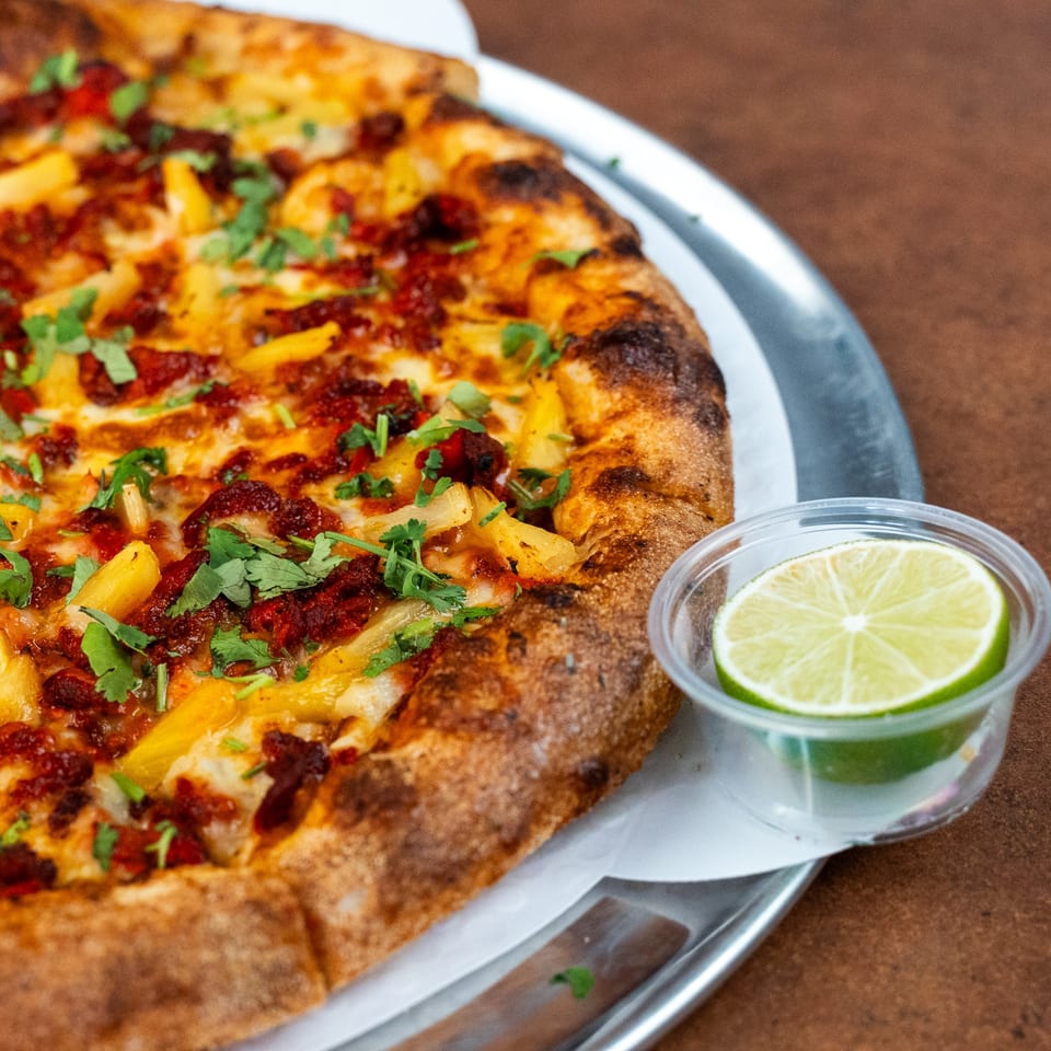A Pizza Buzz pizza with pineapple, chorizo, and cilantro, served with a lime wedge.