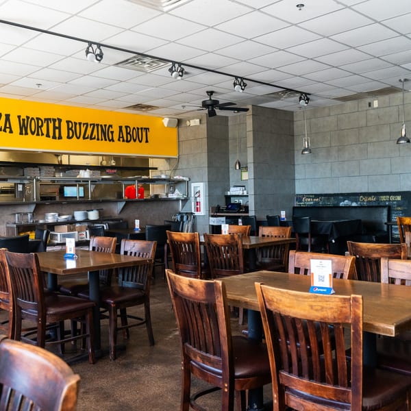 Interior of Pizza Buzz restaurant with empty wooden tables and chairs, and a kitchen area in the background.