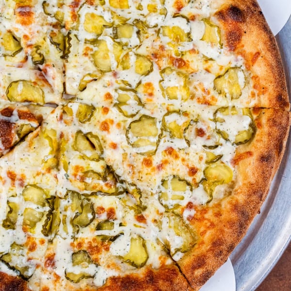 Close-up of a Pizza Buzz pizza topped with sliced pickles, white sauce, and melted cheese.