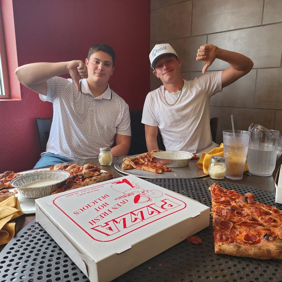 Two young men giving thumbs down to a table full of Pizza Buzz pepperoni pizza slices and a pizza box.