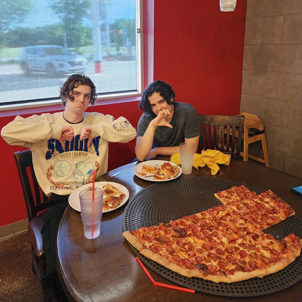 Two young men at Pizza Buzz with a large pepperoni pizza, one giving a thumbs down.