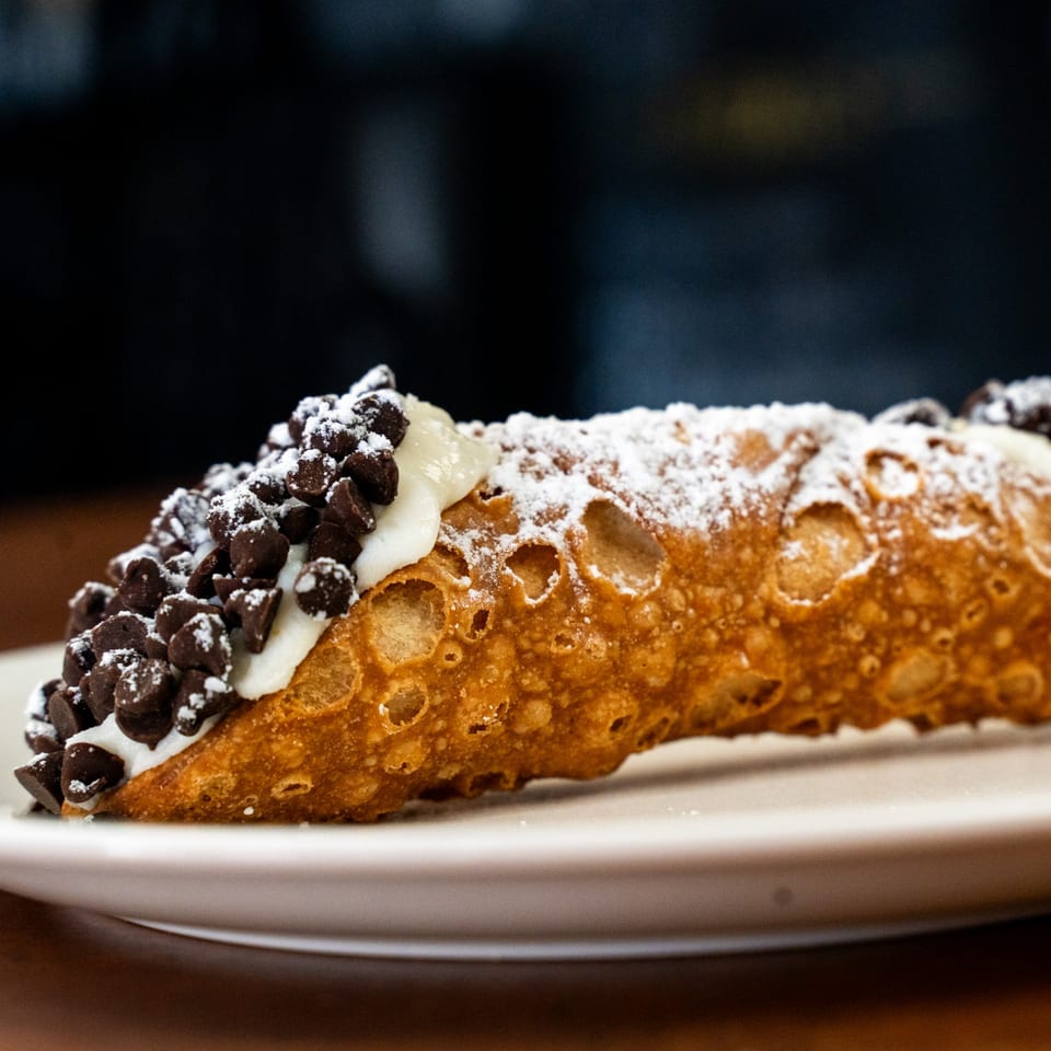 A golden-brown cannoli filled with white cream, topped with chocolate chips and powdered sugar.