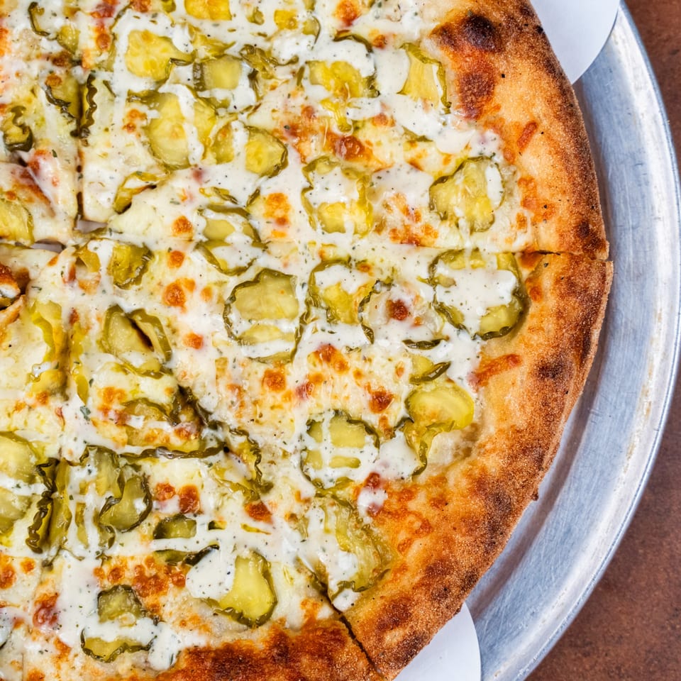 Close-up of a Pizza Buzz pizza topped with white sauce, melted cheese, and sliced pickles.
